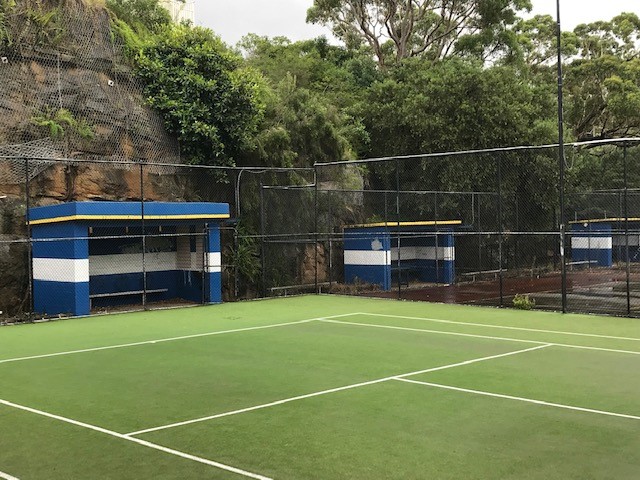 Photo of a green tennis court with fencing, shelters and a rocky cliff face with vegetation in the background 