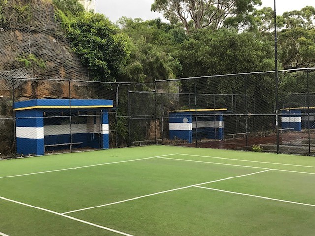 Photo of a green tennis court with fencing, shelters and a rocky cliff face with vegetation in the background