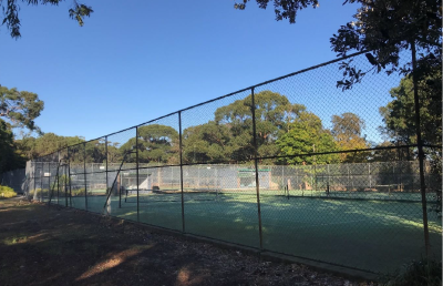 Photo of a tennis court from outside an old and run down fence, taken on a sunny day with trees around the court