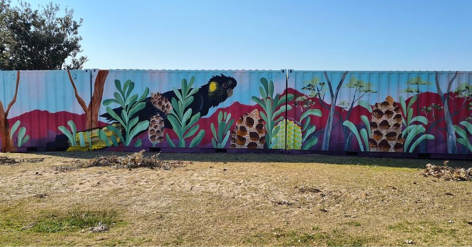 It's a sunny day with clear skies. Sitting on the grass are 3 shipping containers. These containers have been painted with pictures of bright, vibrant vegitation... leaves, flowers and hills in the background. The painting has transformed an eyesore into a pleasant view.